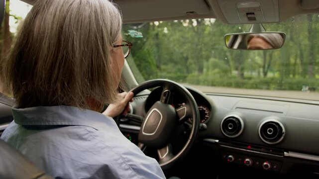 View from behind back rear Caucasian senior old grey haired business woman driving car automobile in city looking around mature aged retired female drive auto grandmother businesswoman taxi driver