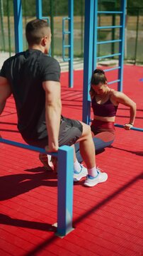 A man and woman perform parallel bar dips during an intense outdoor workout session. The athletic duo is seen exercising together at a modern sports ground under the bright sun.