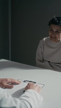 Middle aged Caucasian female psychiatric patient in straitjacket shouting and laughing maniacally during evaluation with senior doctor. Footage is suitable for mental health documentaries, vertical