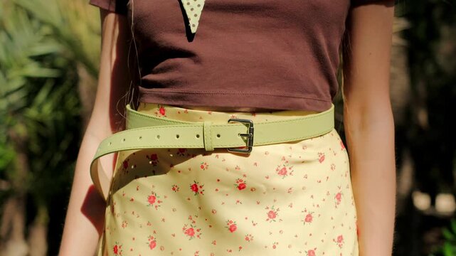 Woman with manicured hands and rings fastening a green leather belt on a yellow floral skirt outdoors on a sunny day