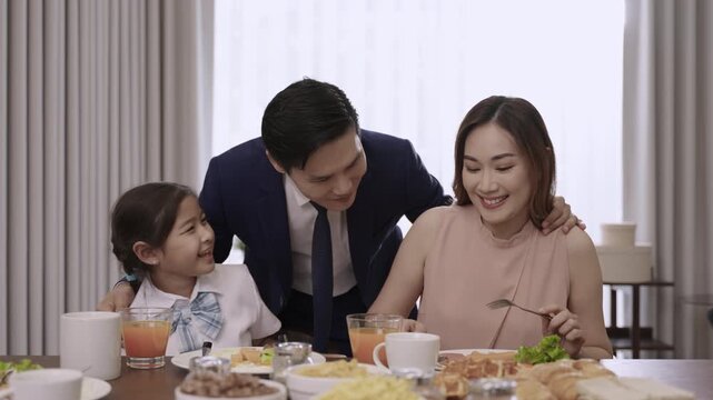 Happy Asian family relationships at the dining table in the morning, with father in a suit, mother, and a child girl smiling, teasing, and bonding before eating breakfast together in kitchen at home. 