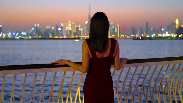 A beautiful woman in a red evening dress standing at Creek Harbour and looking at the distant skyline of Dubai, UAE, during dusk