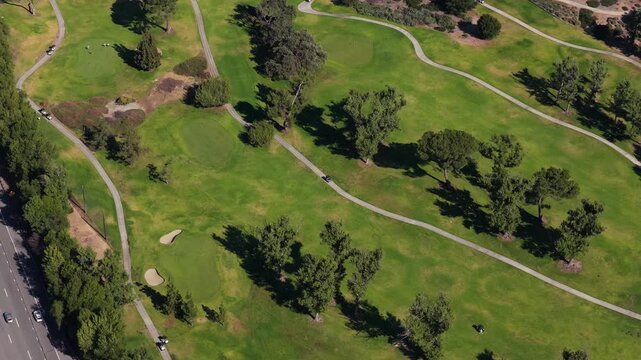 Top down showing a golf course in California, manicured greens, lush fairways, sandy bunkers, winding cart paths, adjacent road traffic