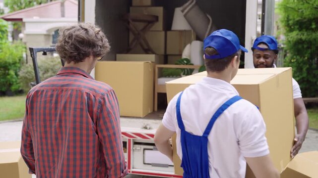 Rear view of a happy young Caucasian man standing, looking, and checking with professional movers in uniform unloading items and furniture from a truck for moving and delivery into the new home. 