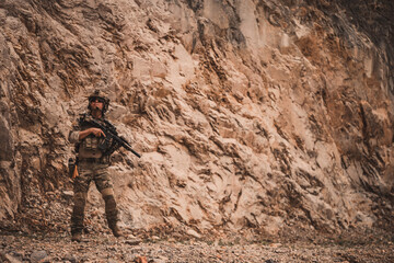 Obraz premium Armed military operator advancing through rugged rocky terrain while aiming a rifle during a combat patrol mission. Tactical soldier moving forward in dangerous combat zone concept.