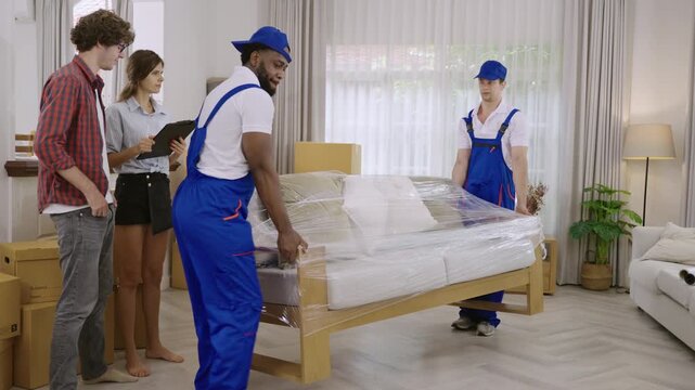 Two professional movers in uniform are moving furniture and cardboard boxes during a home relocation, while a young couple of homeowners, holding a clipboard, are checking for delivery at their home. 