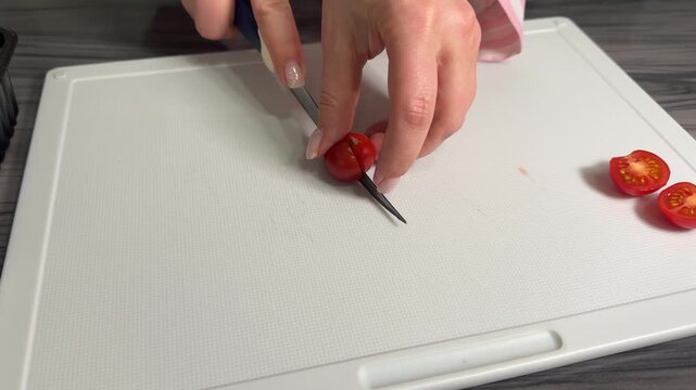 Woman's hands halve cherry tomatoes with a sharp knife on a cutting board, vibrant red juices bursting from fresh, glossy skins in precise slices.