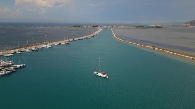 Smooth drone footage performing a partial orbit around a sailing yacht in the calm turquoise waters of Lefkada Marina, Greece. The camera gradually reveals the marina entrance, breakwaters, moored yac