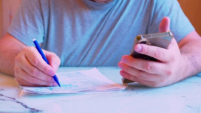 Close-up of a man's hands holding a smartphone and filling out a paper document with a marker.Cinematic shot of cross-referencing data on a cellphone screen and writing on a form in a home office.