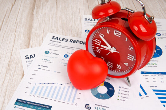 Red heart and alarm clock placed on a sales report dashboard, representing work life balance, time management, productivity, business performance, employee wellbeing, and workplace health concepts.
