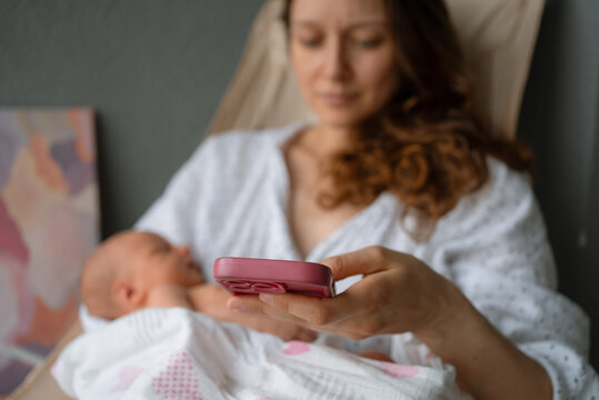 Mother cradling a newborn baby in one arm, actively using a smartphone with the other hand, depicting modern parenthood, multitasking, and digital connectivity in the postpartum journey
