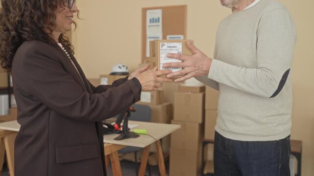 Man carefully hands a cardboard parcel with shipping label to woman over a desk in a small office building stacked with boxes; trust teamwork small business.