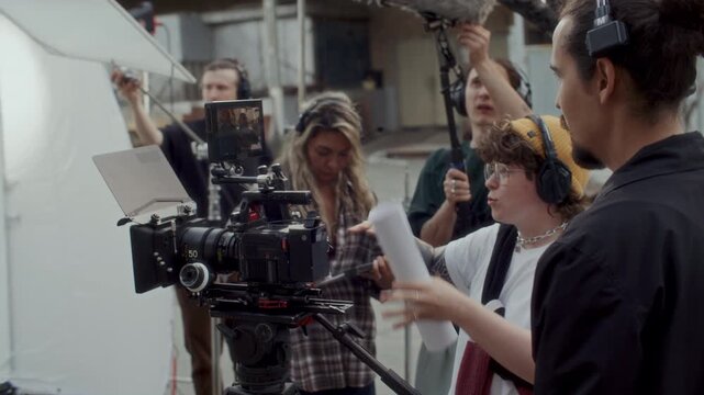 Behind the scenes view of female film director explaining shot composition to camera operator while crew setting up equipment in background during video production shoot on outdoor set