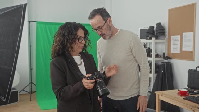 Woman showing camera settings to man in studio while reviewing images on the display and adjusting the lens; collaboration concentration.