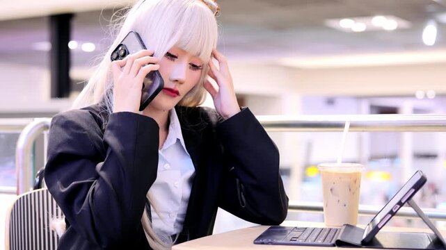 Young woman with blonde hair business attire talking smartphone while using tablet and keyboard, holding coffee cup indoor casual modern cafe focused digital communication technology office work