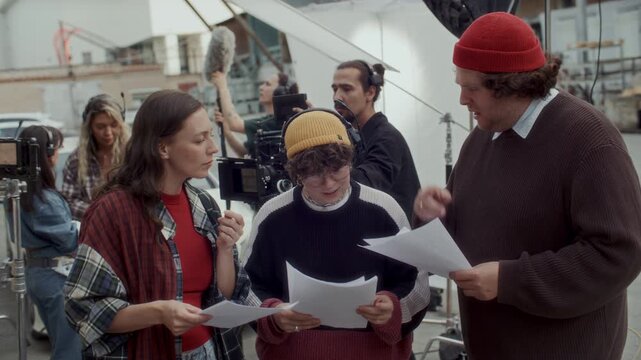 Behind the scenes shot of cast members and female film director reading script pages and discussing dialogue lines, creative team setting up equipment for movie shoot on outdoor urban set