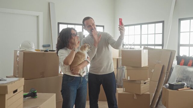 Middle aged couple holding dog and taking selfie amid packed moving boxes inside building; new home happiness.