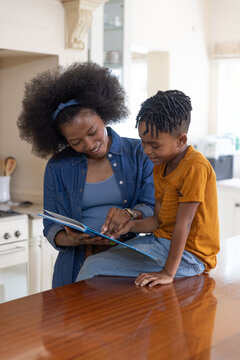 African American mother and son sitting at polished wooden kitchen counter reading picture book