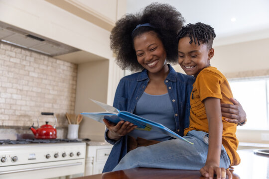 Mother wearing denim shirt and headband sitting with child on kitchen counter, reading picture book