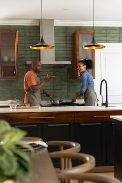 African American couple cooking in kitchen on island wearing aprons using frying pan and spatula