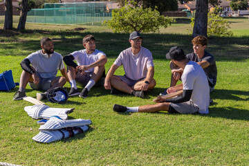 Diverse male teammates sitting on grass at sports field with cricket pads, helmet, bats and bag © wavebreak3