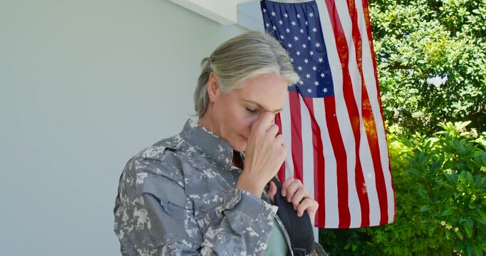 Mid-adult woman in camo steadying pack strap, rubbing nose reflecting on porch beside American flag