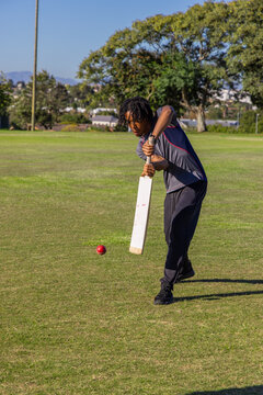 White cricket bat is striking red ball on short grass pitch under clear blue sky