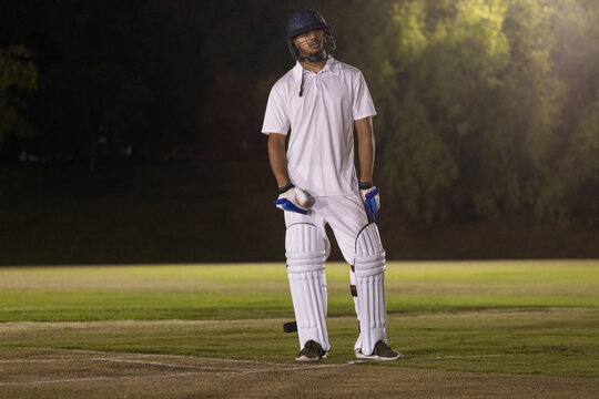 Cricket helmet and bat are resting on floodlit grassy pitch at night near crease lines