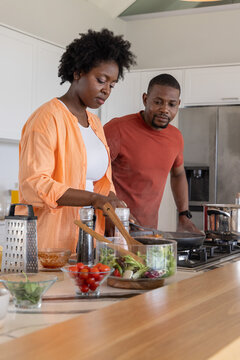 African American couple cooking together, stirring frying pan at kitchen island with salad bowl