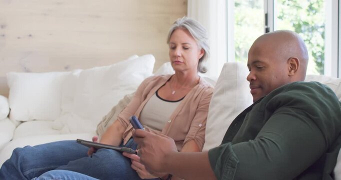 Couple sitting on sofa in living room, man showing phone while woman holding tablet studying screen