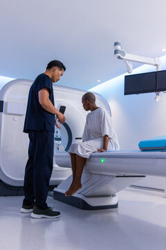 Asian tech in scrubs with remote speaking to African American gowned patient sitting at CT scanner