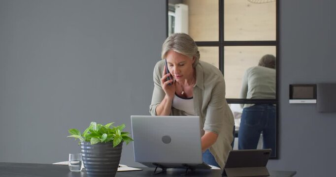Senior woman receiving smartphone call while consulting laptop and writing notes at home workspace