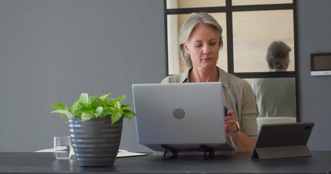 Middle-aged woman glancing at tablet, checking smartphone, answering work call while using laptop