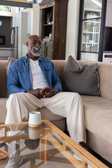 Obraz premium Senior African American man sitting on sofa holding smartphone in denim shirt in living room