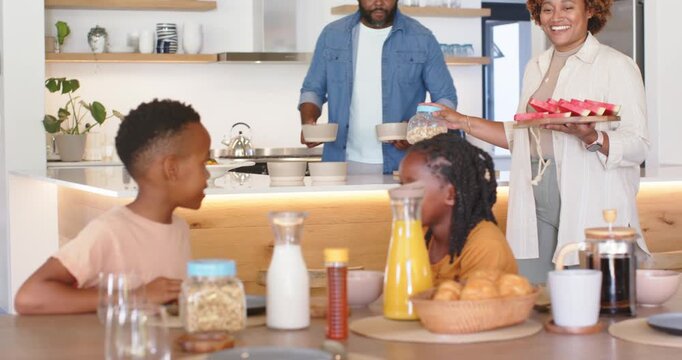 African family mother entering at table carrying jar of cereal and tray, offering meal to children