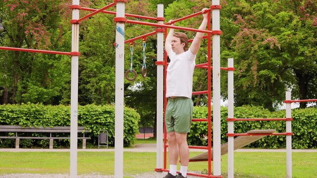 Fit young man doing hanging leg raises on a bar at an outdoor sports ground. Athletic guy training abs and core at a calisthenics park workout. High quality 4k footage
