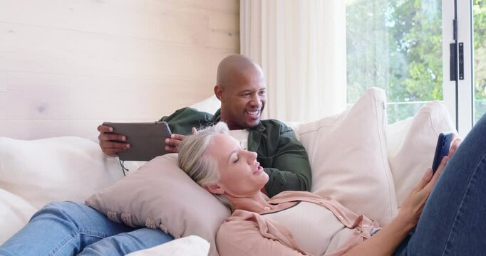 Diverse couple lounging on sofa, woman showing blue phone, man shifting gaze from tablet laughing