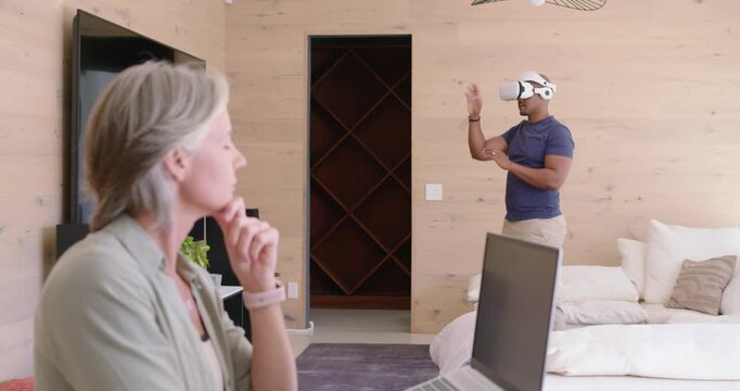 Diverse couple at home, man putting on white VR headset gesturing to interact, woman working laptop