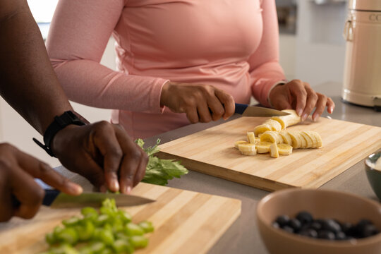 African American couple slicing banana and chopping celery at kitchen counter with knives, berries