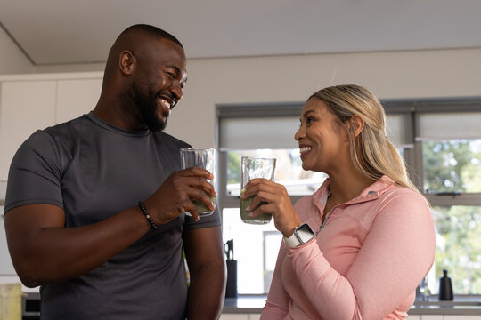African American couple holding green smoothies at kitchen counter, smiling and showing smartwatch