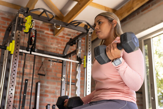 Middle-aged African woman in pink top lifting hex dumbbells on bench in home gym, copy space
