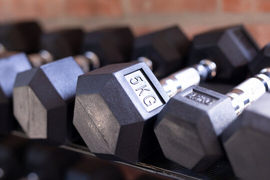 5KG hexagonal dumbbell resting on metal rack in foreground of gym with brick wall backdrop