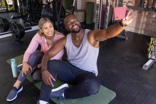 African American friends in sportswear sitting on green mat in gym, taking selfie with pink phone