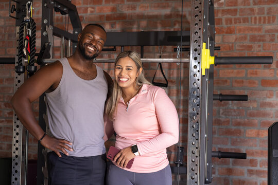 African American couple in workout clothes holding pink smartphone standing by squat rack, barbell