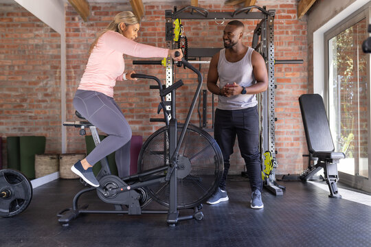 African American coach guiding woman wearing pink top grey leggings pedaling air bike by squat rack