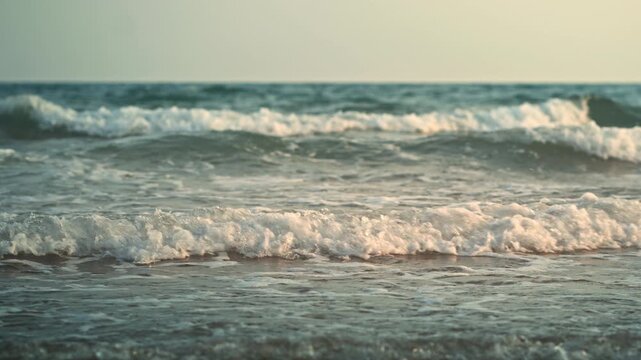 B roll - Close up shot of beautiful sea waves with white foam splashing on the sandy beach shore during sunset. Soft warm sunlight reflecting on the ocean surface, creating a peaceful, calm