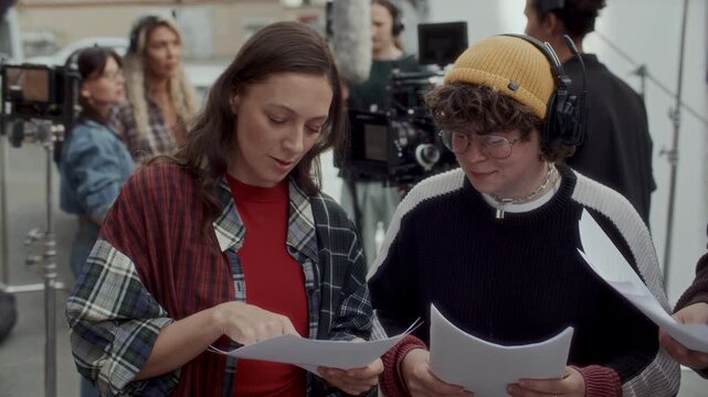 Actors reviewing screenplay and rehearsing dialogue scene with script supervisor during pre-shoot preparations on urban location, professional film crew collaborating in background