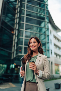 Professional business woman walking in city, carrying laptop and coffee, smiling looking away
