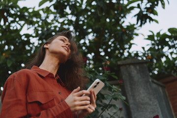 Fototapeta premium A woman stands outdoors in a red shirt, looking up with a phone in hand. Natural light, candid mood, everyday lifestyle moment suggesting curiosity and connection