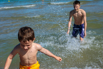Young child running toward camera while teenage boy stands behind in sea water, large water spray in air between them © Татьяна Швец